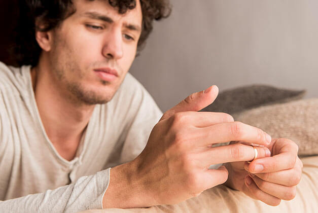 Reasons Why Men Pull Away - Guy on sofa removing wedding ring from finger Reasons Why Men Pull Away - Guy on sofa removing wedding ring from finger