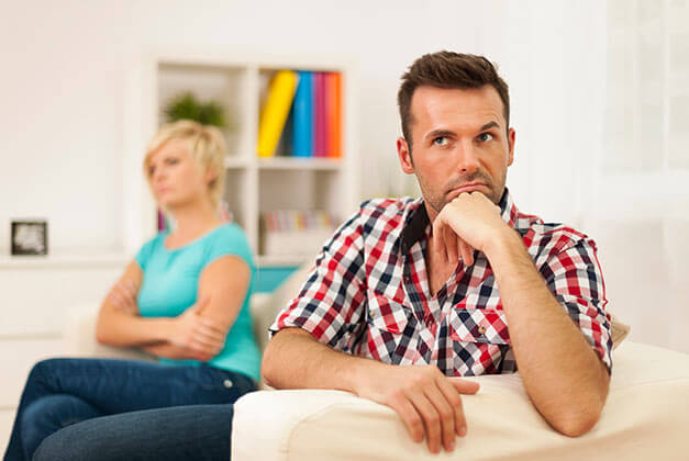 Reasons Why Men Pull Away - Unhappy man thinking with hand on chin and sad girlfriend in background Reasons Why Men Pull Away - Unhappy man thinking with hand on chin and sad girlfriend in background