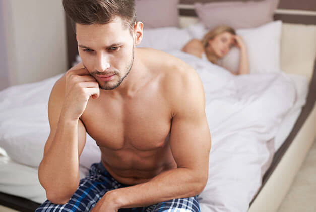 Why Men Pull Away - Sexually unsatisfied man sitting on edge of bed with girlfriend in the background Why Men Pull Away - Sexually unsatisfied man sitting on edge of bed with girlfriend in the background