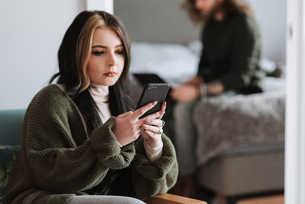 Women That Cheat - Young lady looking at smartphone with her partner sitting on bed in background Women That Cheat - Young lady looking at smartphone with her partner sitting on bed in background